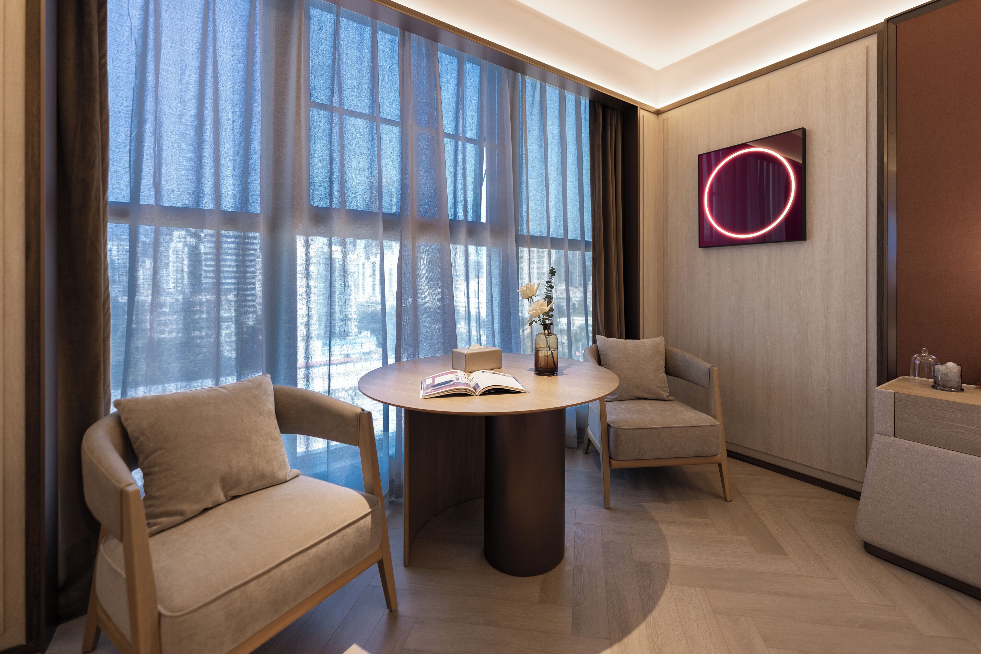 A modern room with a round table, two chairs, a bookshelf, and a large window with curtains.