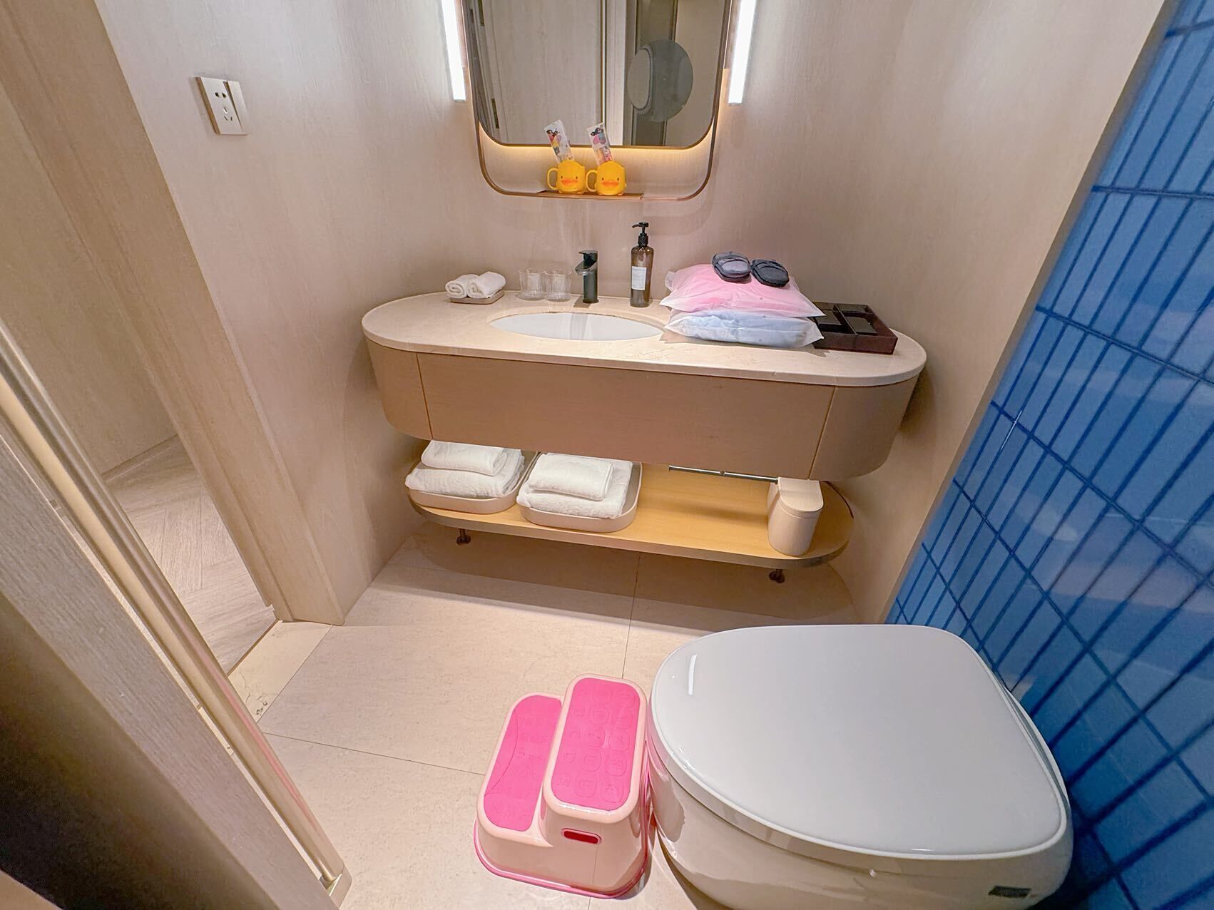 Un baño con inodoro, lavamanos, espejo y repisas con toallas.