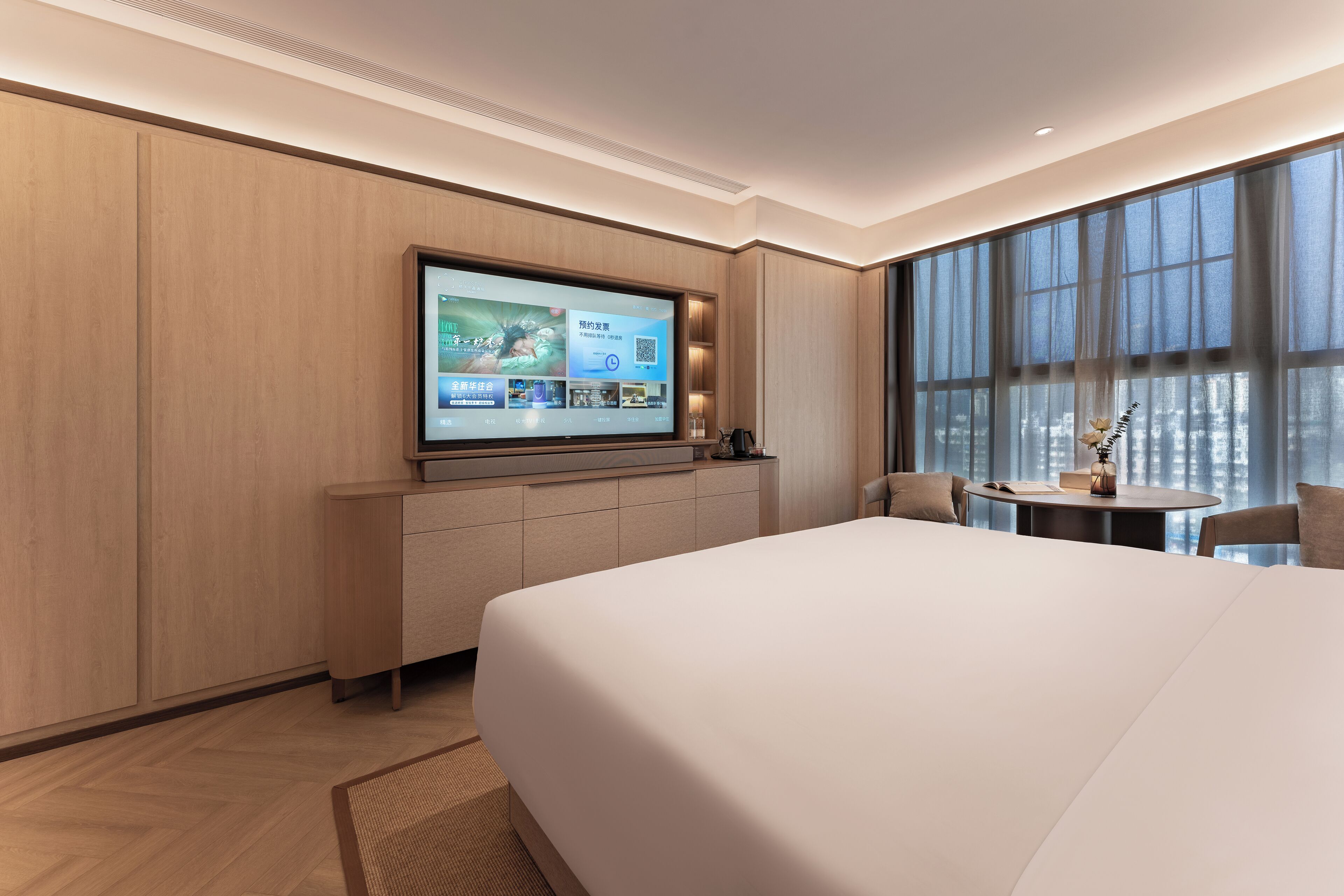 A hotel room with a large bed, a TV mounted on the wall, a small table with a chair, and a window with curtains.