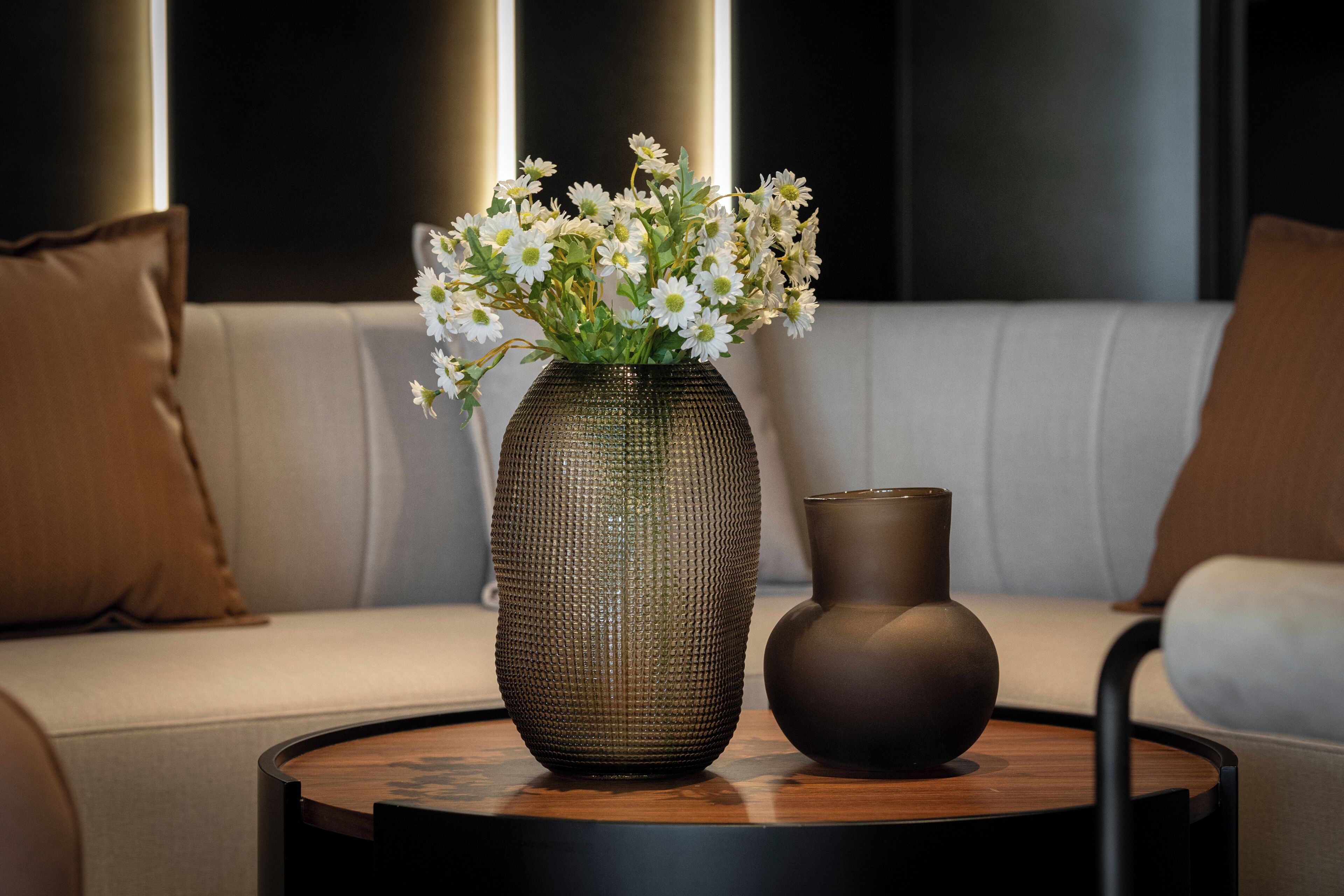 Una mesa redonda de madera con dos jarrones, uno con flores blancas y otro vacío, sobre un sofá con cojines marrones.