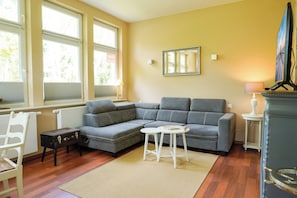 Ein Wohnzimmer mit einem Ecksofa, einem kleinen Tisch, einem weißen Sessel, einem Kamin und großen Fenstern.