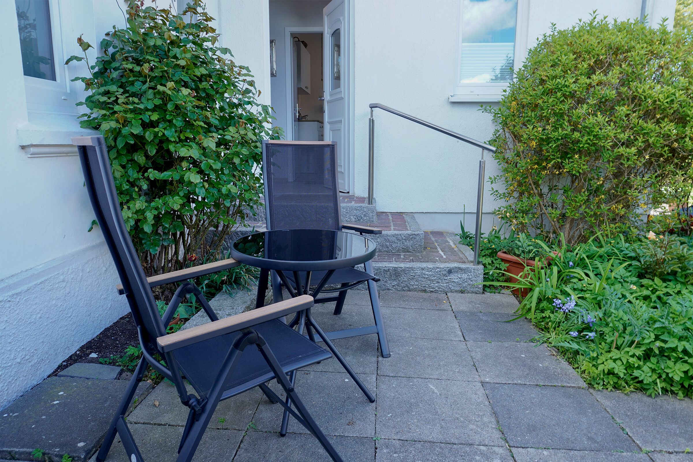 Eine Terrasse mit zwei Stühlen und einem Tisch, ein weißes Gebäude und ein grüner Busch.