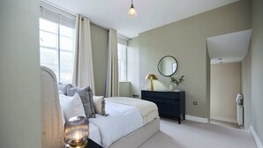 Une chambre à coucher comprenant un lit, une table de chevet, une lampe, une commode, un miroir et une fenêtre avec des rideaux.