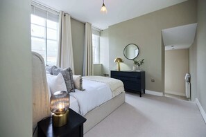 Ein Schlafzimmer mit Bett, Nachttisch, Lampe, Kommode, Spiegel und einem Fenster mit Vorhängen.
