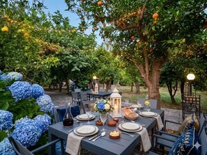 An outdoor dining area with tables set for a meal, surrounded by trees and blue hydrangea bushes. - Meta CC Palace  (Meta)