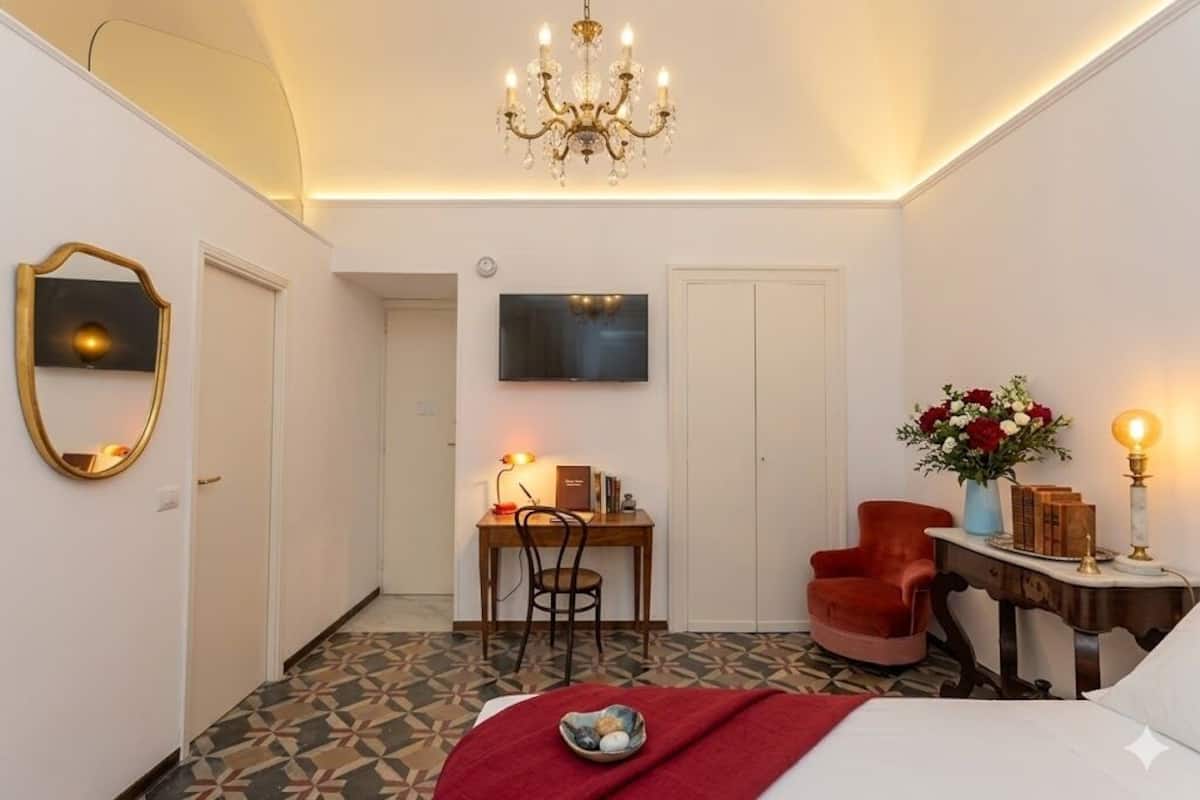 Ett hotellrum med en säng, ett skrivbord med en stol, en tv, en röd fåtölj, ett litet bord med en lampa och en kristallkrona.