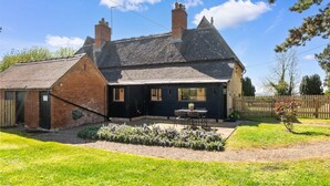 A two-story house with a brick exterior, a tiled roof, and a small garden with a patio table and chairs. - Alms House West, Grade II Listed, Garden & Patio (Forthampton)