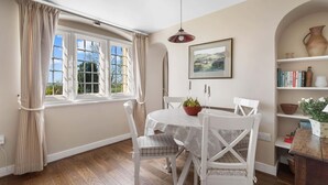 A dining area with a table, chairs, a pendant light, a window with curtains, a bookshelf, and a painting. - Alms House West, Grade II Listed, Garden & Patio (Forthampton)