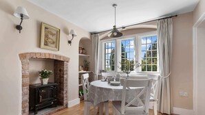 Un coin repas avec une cheminée en briques, des chaises blanches, une table ronde et une grande fenêtre avec des rideaux.