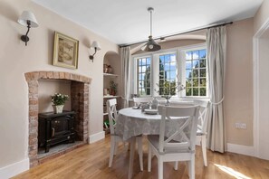 A dining area with a brick fireplace, white chairs, a round table, and a large window with curtains. - Alms House East, Grade II Listed, Garden & Patio (Forthampton)