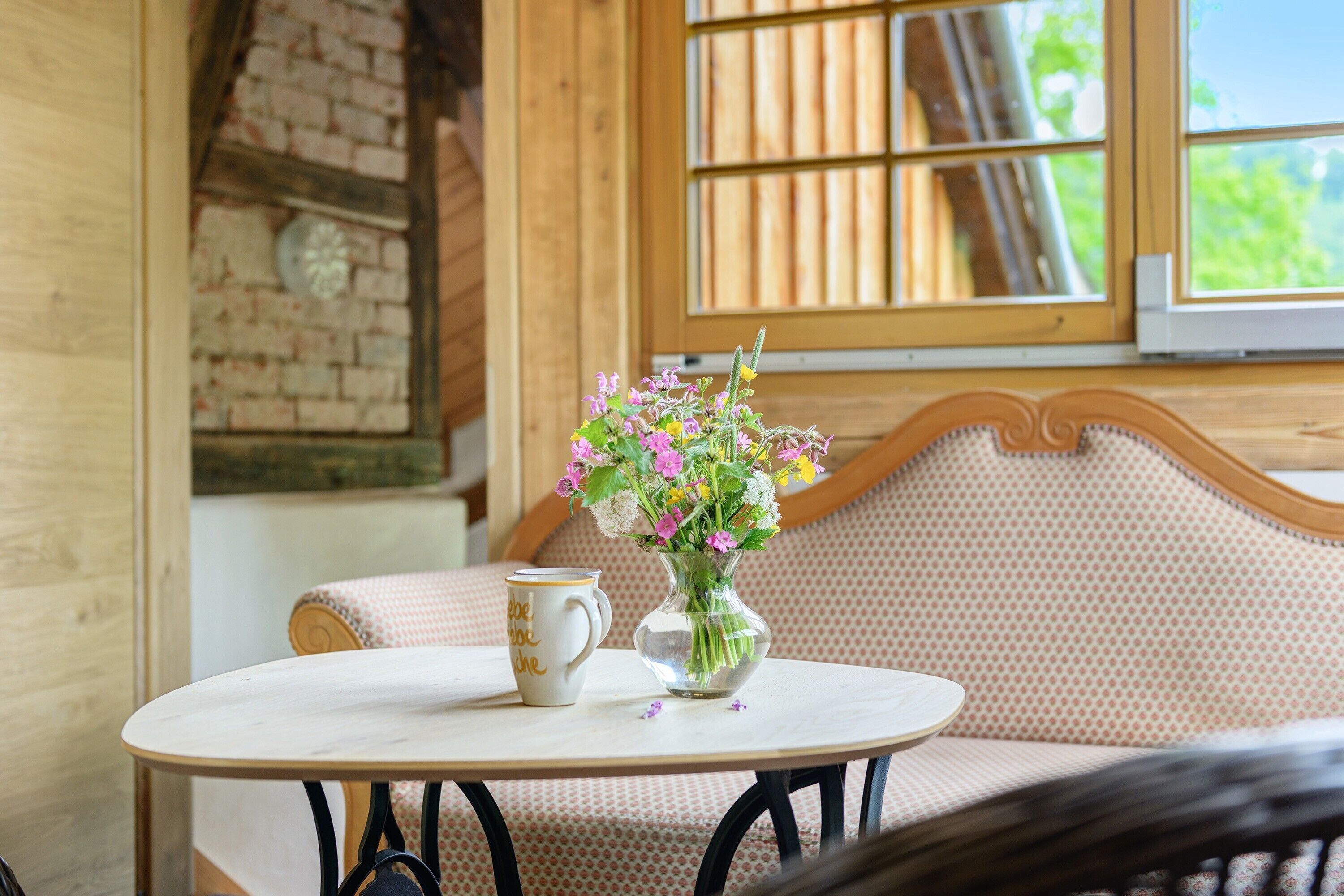 A round table with a white mug and a clear vase with flowers, set against a wooden wall and a patterned sofa.