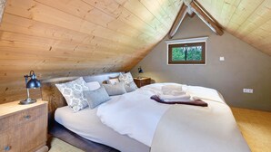 Una camera da letto con soffitto in legno, un letto con biancheria bianca, un comodino con una lampada e una finestra vista alberi.