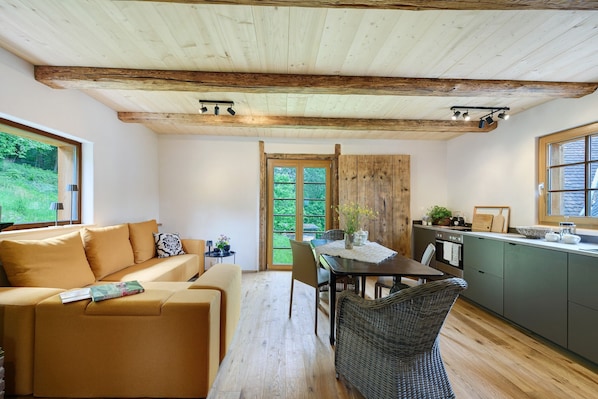 Ein gemütliches Interieur mit Holzbalkendecke, einer Essecke mit Tisch und Stühlen, einer Küche mit grünen Schränken und einem großen Fenster mit Blick ins Grüne.