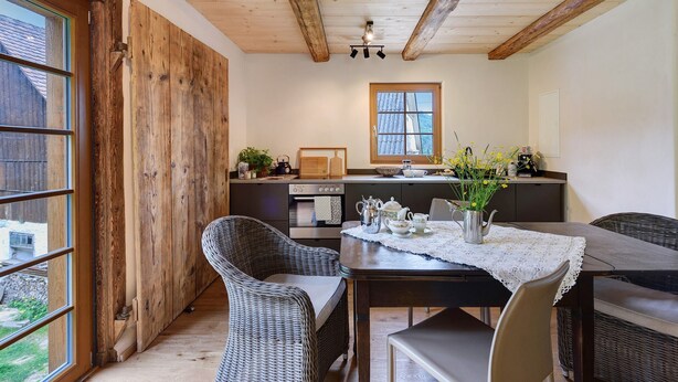 A dining area with wicker chairs, a table with a lace tablecloth, and a view of the kitchen in the background.