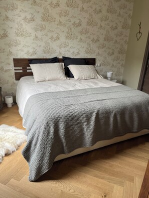 Ein ordentlich bezogenes Bett mit einer grauen Strickdecke, einem hölzernen Kopfteil und einer gemusterten Tapete.