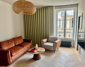 Ein Wohnzimmer mit braunem Sofa, einem beigen Sessel, einem runden Couchtisch, einer gewobenen Pendelleuchte und Blick auf Gebäude durch ein Fenster mit grünen Vorhängen.