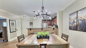 Un coin repas avec une table en bois, des chaises, un lustre et une cuisine en arrière-plan.