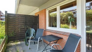 A patio with two chairs and a table, a wooden fence, and a window with a view of greenery. - Apartment 'In The House Juist' with Private Terrace and Wi-Fi (Dornum)