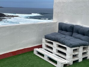 Ein Balkon mit einer grauen Sitzgelegenheit mit Polstern, einer Betonwand und Blick auf das Meer.