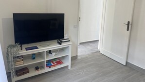 Huoneessa on valkoinen, litteänäyttöinen televisio, joka on sijoitettu valkoiselle jalustalle. Huoneessa on myös suljettu ovi ja valkoinen hylly, jossa on kirjoja ja kaukosäädin.