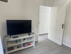 Ein Zimmer mit einem Flachbildfernseher auf einem weißen Standfuß, einer geschlossenen Tür und einem weißen Regal mit Büchern und einer Fernbedienung.
