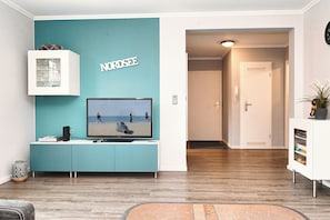 Ein Wohnzimmer mit einer türkisfarbenen Akzentwand, einem Fernseher, einem weißen Schrank und Holzboden.