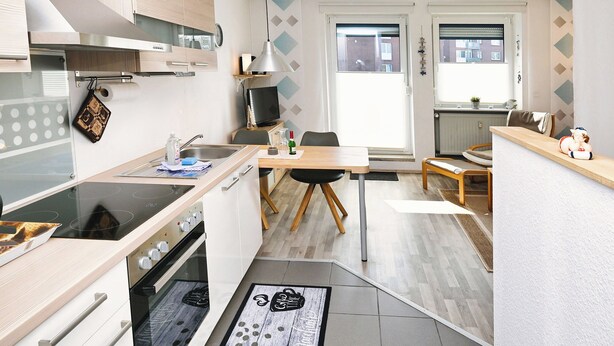 Eine kompakte Küche mit Herd, Backofen und Spüle, ein Essbereich mit Tisch und Stühlen sowie Blick auf die Straße durch ein Fenster.