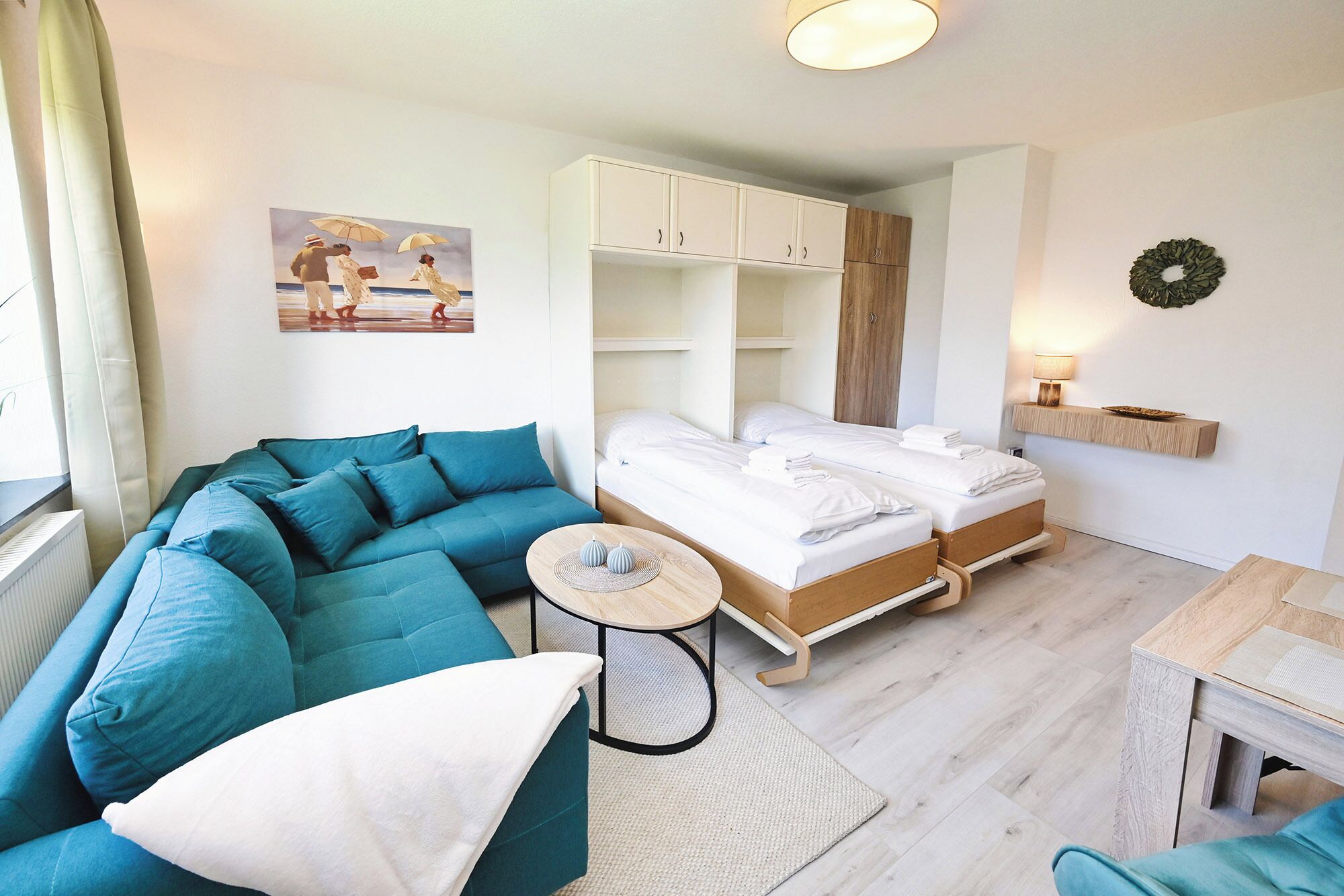 A modern room with a teal sofa, a bed with two white linens, a small round table, and a wooden cabinet.
