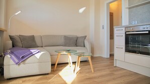 A modern living room with a sofa, a glass coffee table, and a kitchen area with an oven. - Dünentraum 9 in Grömitz: 34 m² with a balcony, private parking, and Wi-Fi (Grömitz)