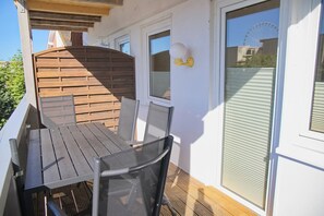 Eine Holzterrasse mit Tisch und Stühlen, ein Fenster mit Blick auf ein Riesenrad und eine weiße Wand mit einer Leuchte.