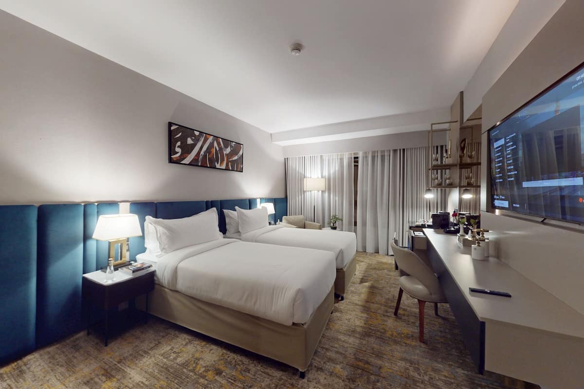 Quarto de hotel com duas camas, uma escrivaninha, televisão e uma janela grande com cortinas.