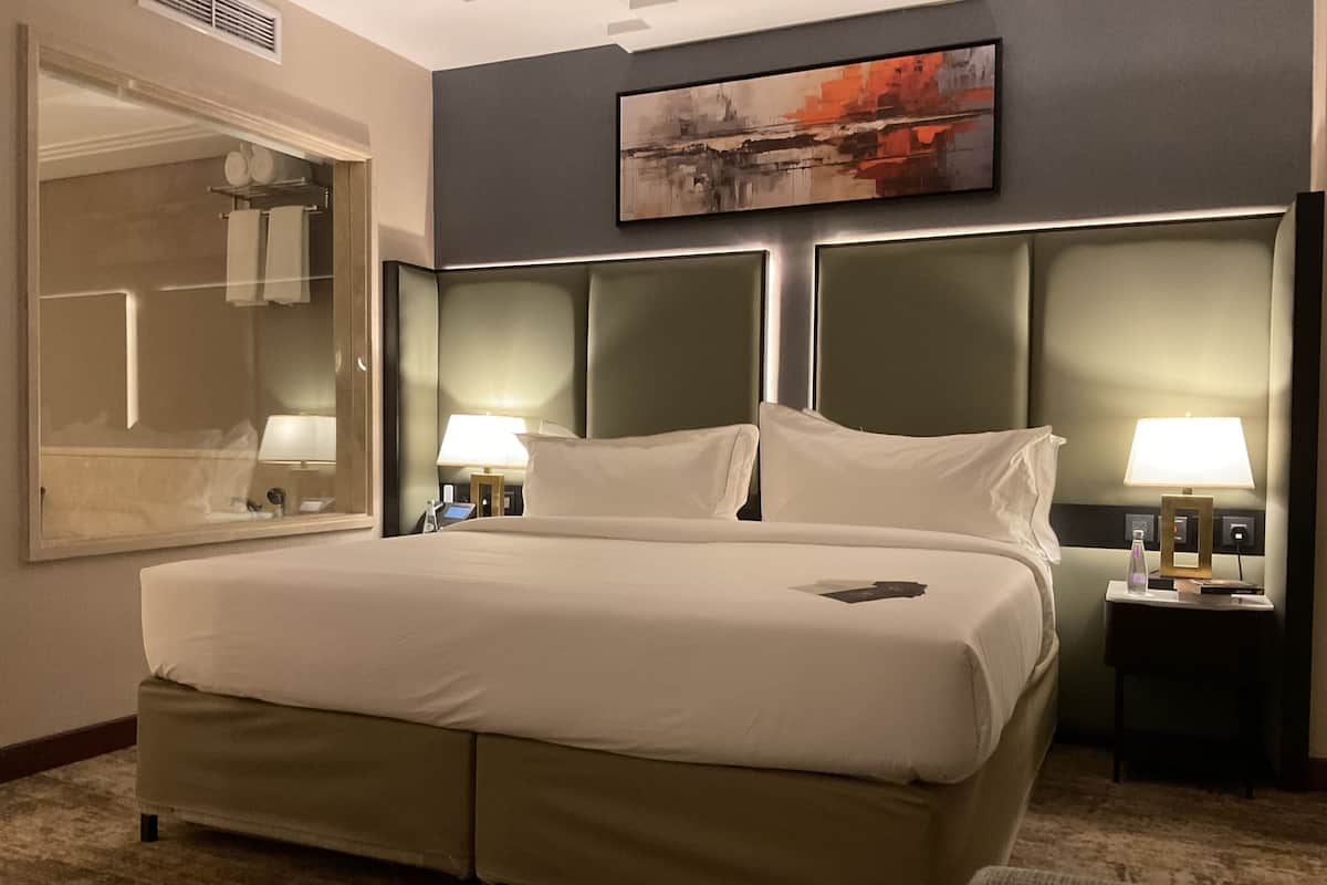 Quarto de hotel com uma cama grande, duas mesas de cabeceira com luminárias, um espelho grande e um quadro na parede.