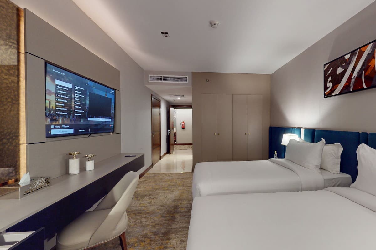 Quarto de hotel com televisão exibindo lista de canais, escrivaninha com cadeira, duas camas, cabeceira azul e uma grande pintura abstrata na parede.