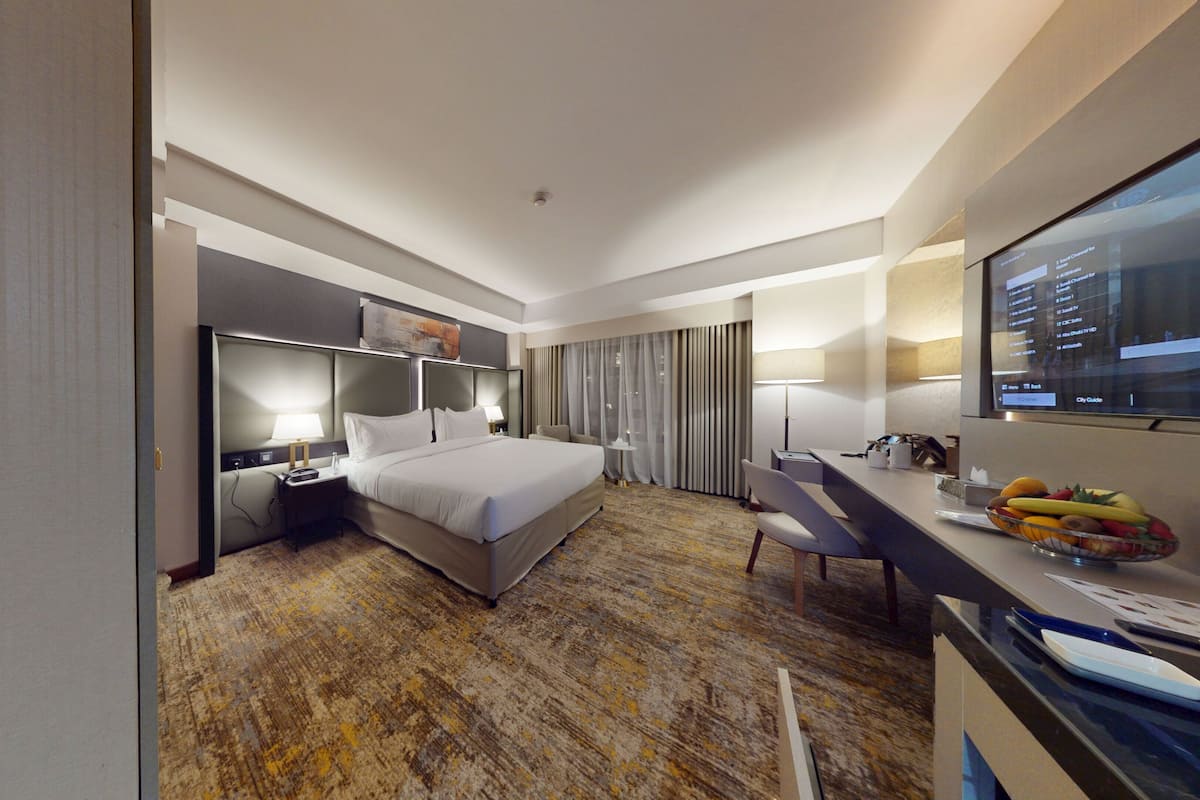 Quarto de hotel com cama, escrivaninha, cadeira, televisão e uma cesta de frutas.