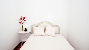Un letto con biancheria bianca, un comodino bianco con una lampada e un vaso con fiori rossi.