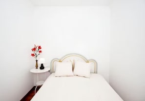Ein Bett mit weißer Bettwäsche, ein Nachttisch in Weiß mit einer Lampe und eine Vase mit roten Blumen.