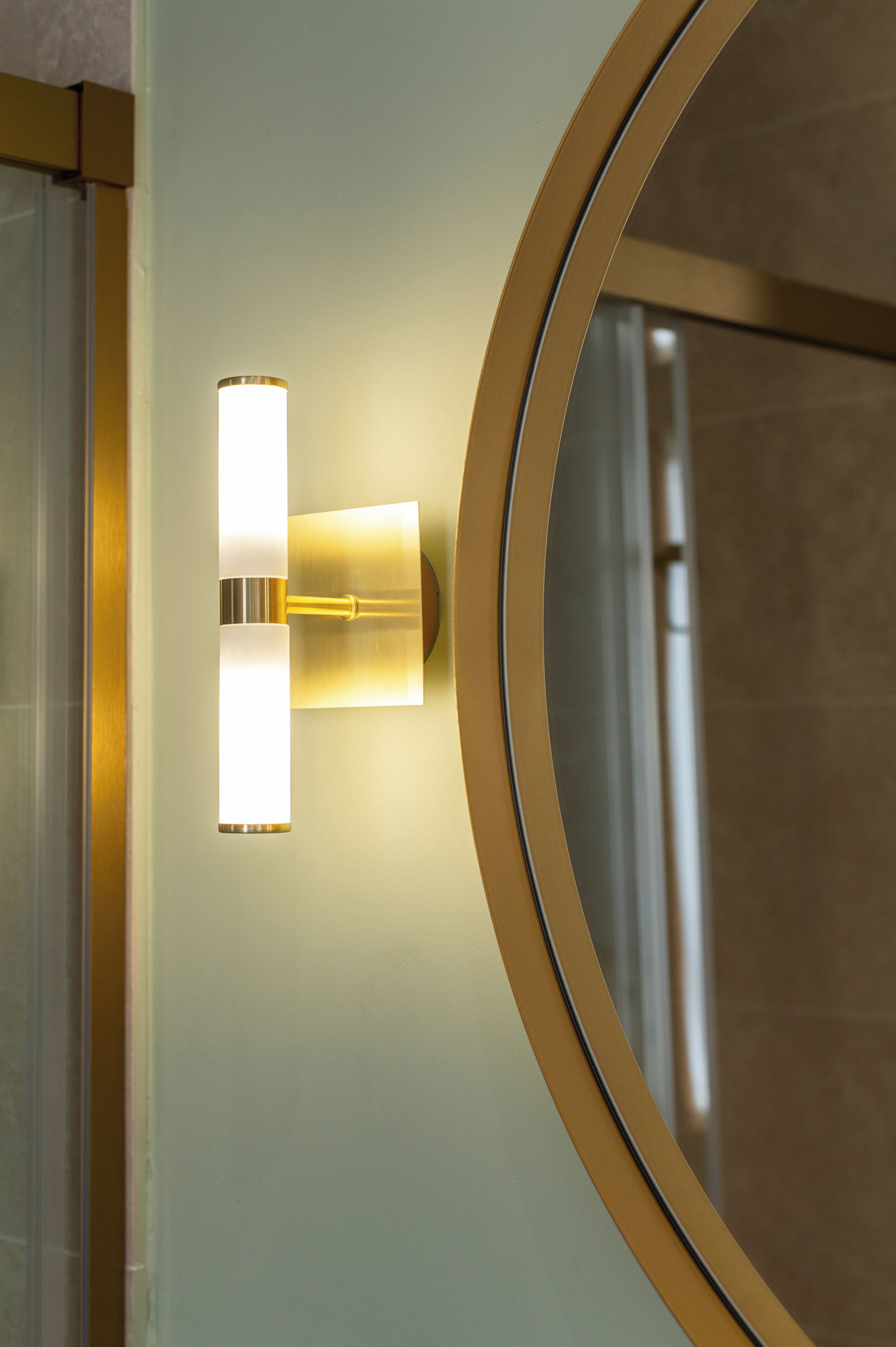 Een wandlamp met een rechthoekige vorm en een cilindrische basis, geplaatst naast een ronde spiegel met een gouden lijst.
