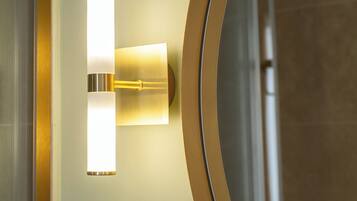 Een wandlamp met een rechthoekige vorm en een cilindrische basis, geplaatst naast een ronde spiegel met een gouden lijst.