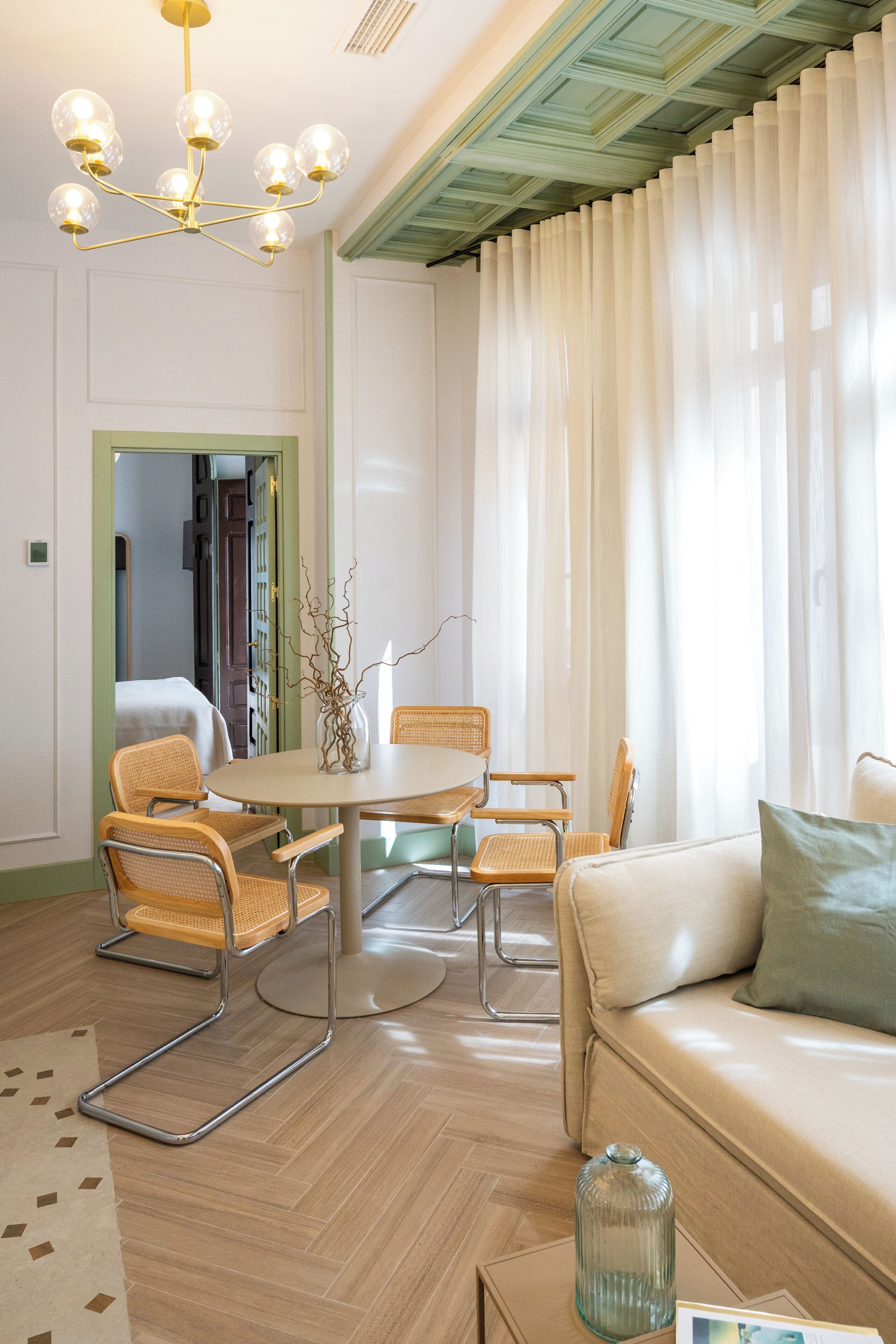 Ein Zimmer mit Esstisch, Stühlen, einer Couch, einer Chandelüstere und einem großen Fenster mit Vorhängen.