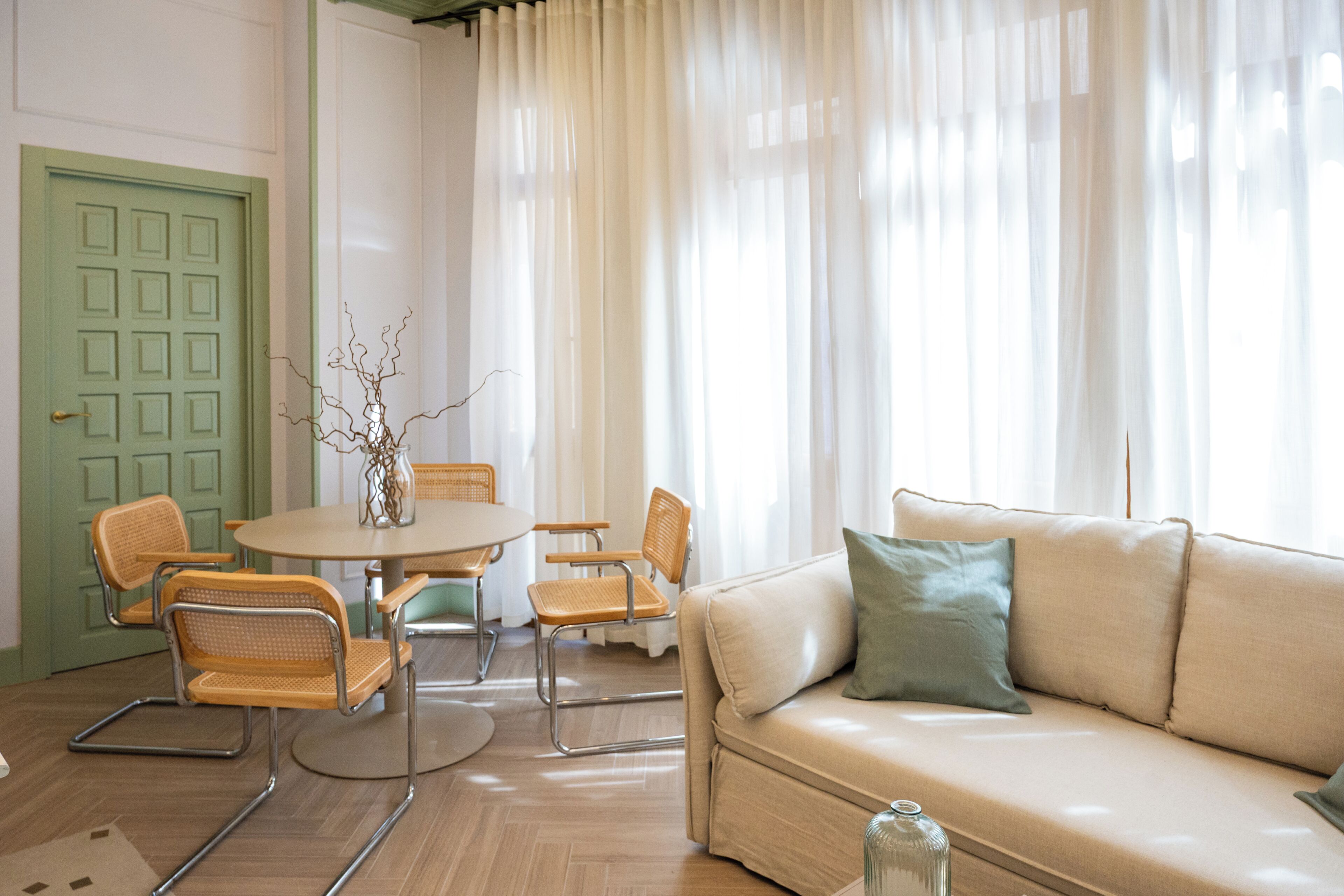 A room with a green door, a round table with chairs, a beige sofa, and a vase with branches.