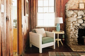 A cozy room with a stone fireplace, a small table with a lamp, and a chair with a cushion.