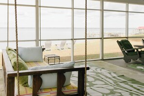Ein hölzerner Schaukelstuhl mit Kissen, ein kleiner Tisch und Blick auf den Strand durch große Fenster.