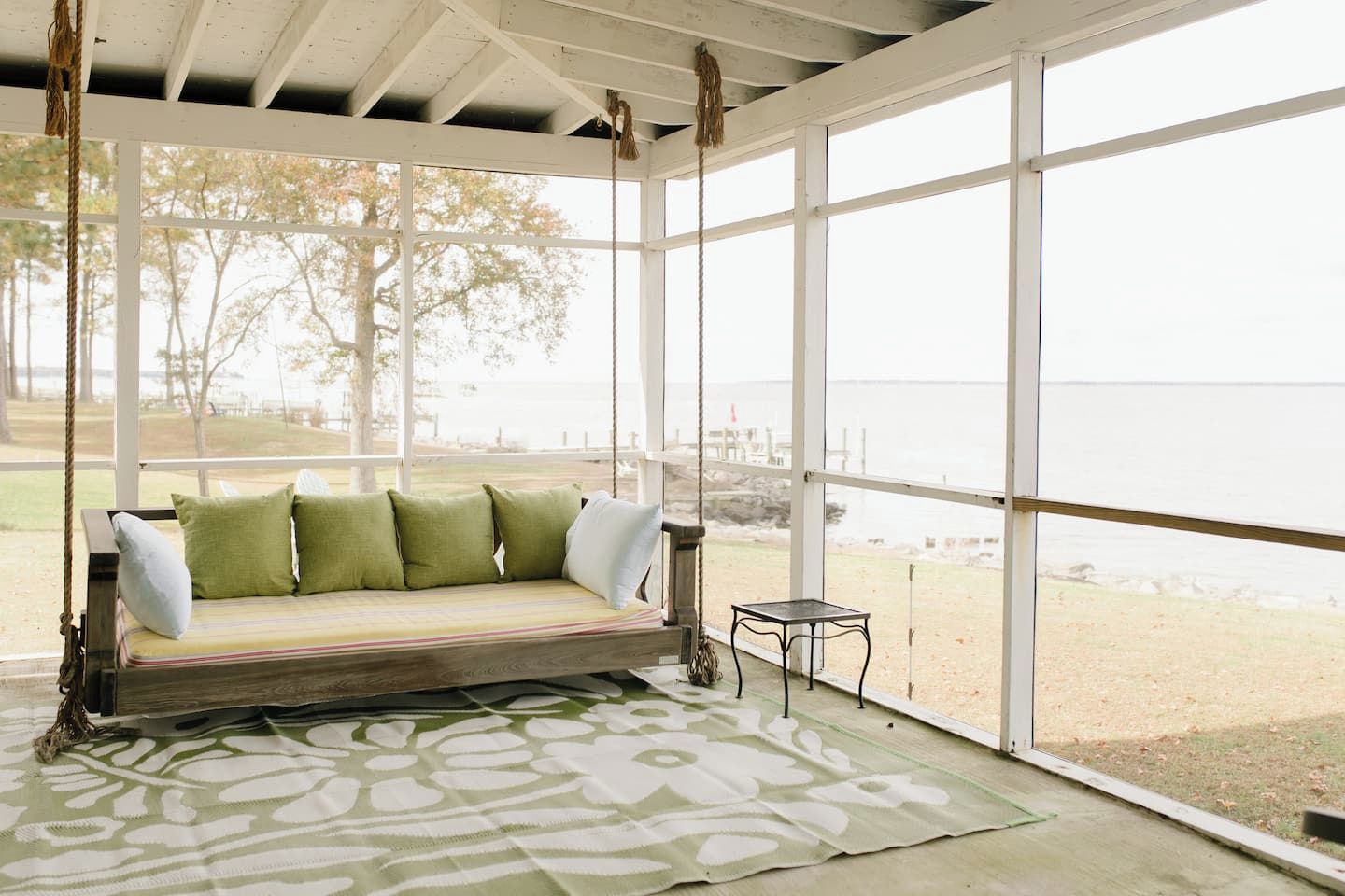 A covered outdoor seating area with a swing seat, a patterned rug, and a view of the beach.