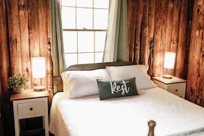 A bedroom with a wooden headboard, a bed with white bedding, a green pillow with 'Rest' text, two bedside tables with lamps, and a window with curtains.