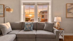 En stue med en grå sofa, en lampe og udsigt til en solnedgang gennem et vindue.