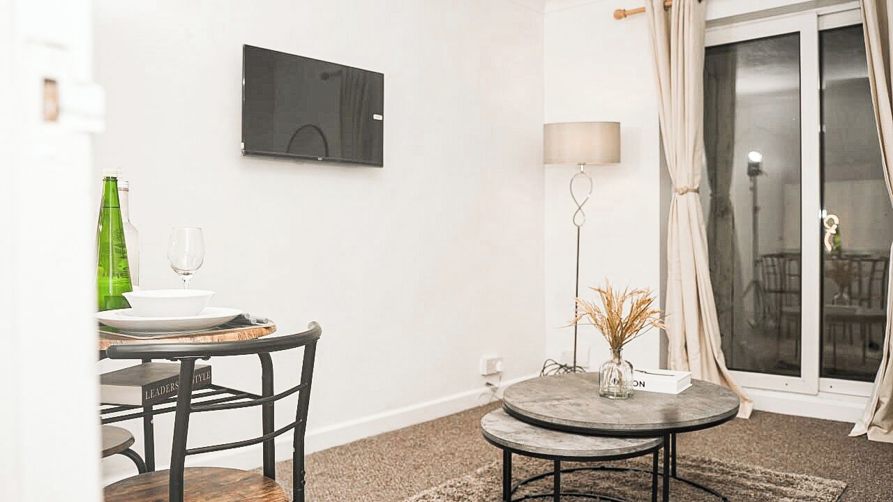 Een eethoek met een tafel, stoelen, een aan de muur bevestigde televisie, een lamp en uitzicht door een glazen deur.