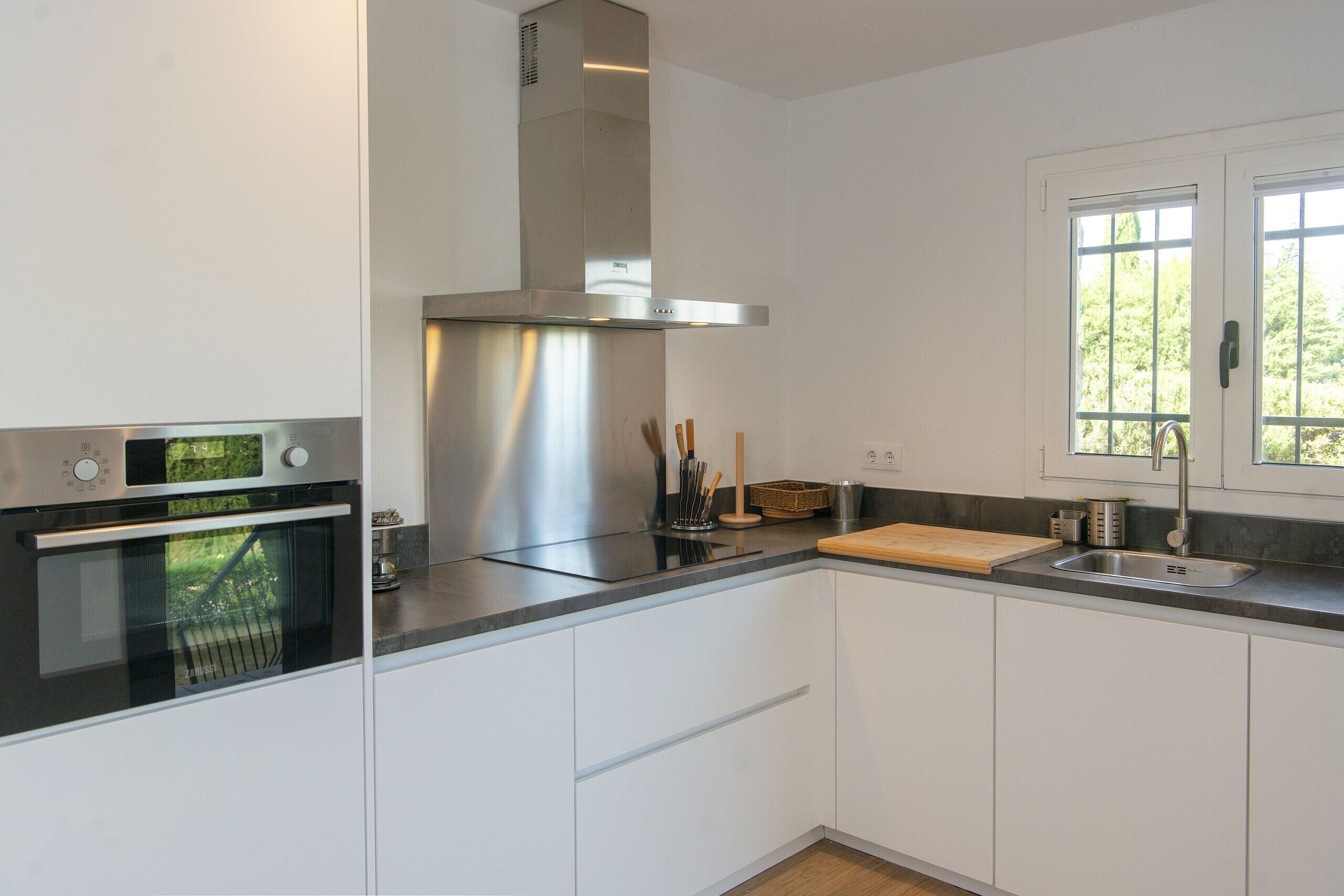 A modern kitchen with white cabinets, a stainless steel range hood, a built-in oven, a wooden cutting board, and a window with a view of greenery.