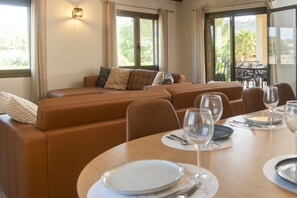 Ein Essbereich mit Tisch für zwei Personen, einer Couch mit Kissen und Blick ins Freie durch große Fenster.
