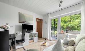 A living room with a TV, a sofa, a dining table, and a large window with a view of greenery. - "Ankerplatz" Vacation Apartment in Büsum: With Terrace, Wi-Fi, Family-Friendly, (Büsum)