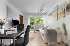 A modern living room with a television, dining table, and sofa, a large window with a view of greenery, and artwork on the walls. - "Ankerplatz" Vacation Apartment in Büsum: With Terrace, Wi-Fi, Family-Friendly, (Büsum)